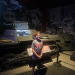 A young boy stands in front of a military tank display at a museum, showcasing historical military vehicles and equipment. Military tank exhibit at museum.