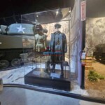 Showcasing a WWII soldier's uniform with a camera operator capturing the display. Historical WWII military uniform exhibit in a glass case at Finish Masters Construction & Painting.