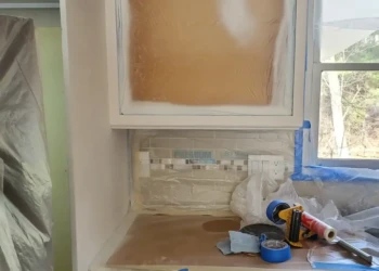 Professional cabinet refinishing doesn't stop after primer is applied. At Finish Masters, every primer coat is allowed to cure, inspected, and sanded smooth before the next coat is applied because our reputation is built on the details others overlook. Finish Masters cabinet refinishing project in Tewksbury Massachusetts showing primed cabinet boxes near the refrigerator and dishwasher protected with masking while the first coat of primer cures before sanding and recoating.