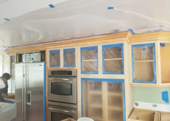 Kitchen cabinets being refinished with masking tape and plastic covering.