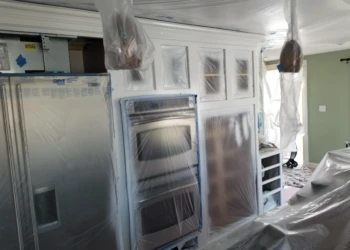 Cabinet refinishing in progress with plastic coverings and painting equipment.