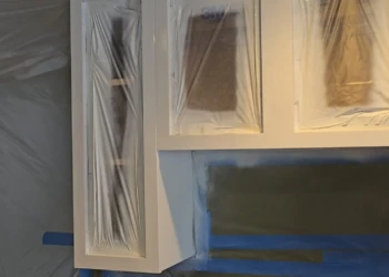 The transformation is officially underway. After extensive preparation, repairs, sanding, masking, and dust containment, the first coat of Benjamin Moore AF-70 Storm begins turning these dated wood cabinets into a modern, furniture-grade finish worthy of the Finish Masters name. Finish Masters cabinet painting project in Burlington Massachusetts showing upper cabinets sprayed with the first coat of Benjamin Moore AF-70 Storm while cabinet interiors remain masked and protected during the finishing process.