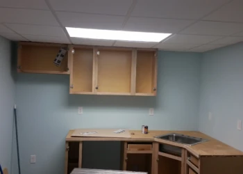 Unfinished oak kitchen cabinets installed in a basement with light blue walls and fluorescent ceiling light
