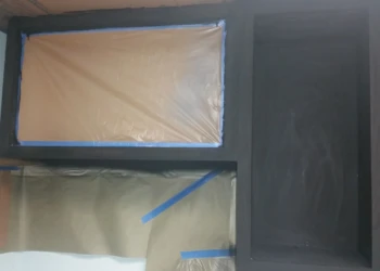 Black oak cabinets with taped and paper-covered sections in a basement kitchen during refinishing work