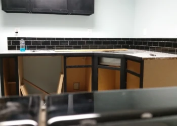 Partially installed black painted oak kitchen cabinets with no doors and black tile backsplash in a basement kitchen