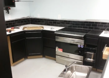Corner of a basement kitchen featuring matte black painted oak cabinets with black subway tile backsplash and stainless steel stove and sink