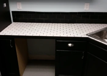 Black painted oak kitchen cabinets below a white tile countertop with small dark grout lines and stainless steel sink