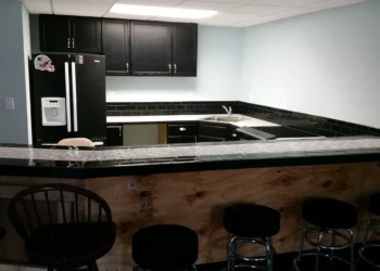 Basement kitchen featuring black painted oak cabinets, black tile countertop, and black refrigerator in Billerica MA
