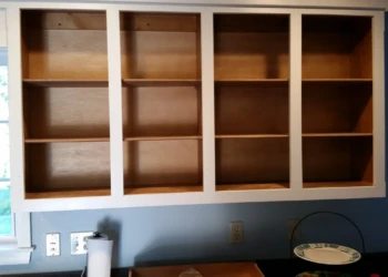 Empty wooden kitchen cabinets with white painted frames mounted on a blue wall by a window