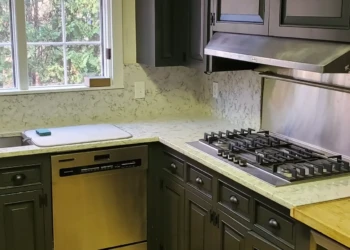 Dark gray painted kitchen cabinets surrounding a stainless steel cooktop and dishwasher with light granite countertops and pine hardwood flooring in Bedford, NH
