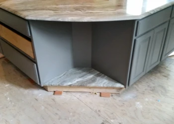 Gray-painted kitchen island with marble countertop and multiple cabinet doors in an unfinished floor setting