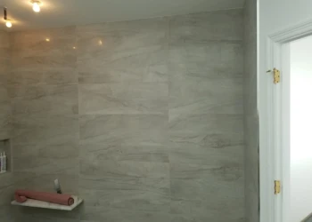 Close-up of large gray marble-effect tiles on a master bathroom wall near a white door with gold hinges in Carlisle MA