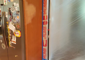 Wood kitchen cabinet next to stainless steel refrigerator with plastic sheeting and painter's tape for cabinet refinishing project in Groton MA