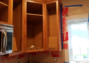 Open wooden kitchen cabinets without doors during cabinet refinishing in Groton, MA, with plastic sheeting protecting walls and window