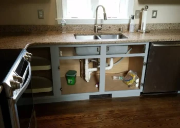 Kitchen cabinets below a granite countertop with open lower cabinet doors showing plumbing pipes beneath the sink in a Groton MA kitchen