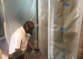 Craftsman wearing protective mask refinishing kitchen cabinet in Groton home with plastic sheeting