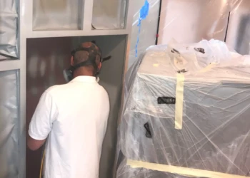 Person spray painting kitchen cabinets inside a home with surfaces and appliances covered for protection