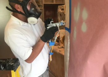 Worker wearing protective mask and gloves spraying a surface during cabinet refinishing in Groton MA