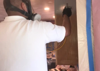 Worker wearing a protective mask and gloves spray-painting kitchen cabinets during refinishing in Groton MA