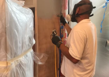 Technician wearing protective gear spraying finish on kitchen cabinets covered in protective plastic in Groton MA