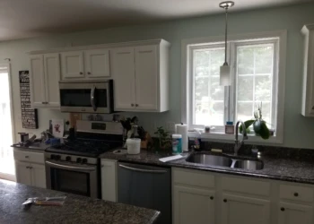 White painted kitchen cabinets with silver hardware below and above granite countertops near a large window in a Merrimack kitchen