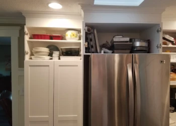 White finished kitchen cabinet with open upper doors showing stored baking pans above stainless steel refrigerator in Nashua kitchen