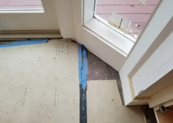 Temporary protective cardboard and tape covering hardwood floor next to door and window frame in renovation site.