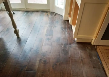 Dark stained hardwood floor in kitchen with scratches and dust near white cabinets and glass door.