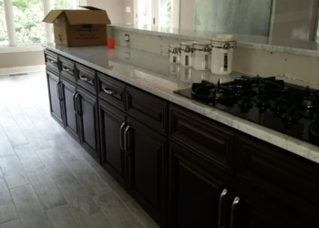 Dark wood kitchen cabinets with silver handles and marble countertop beside a gas stove in a bright kitchen