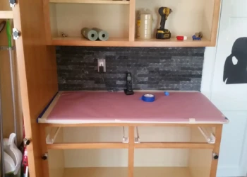 Wood kitchen cabinet frame with protective paper on countertop and tools in open shelving.