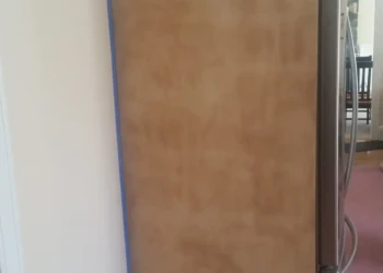 Side panel of a tall wooden kitchen cabinet next to a refrigerator, taped around edges for refinishing preparation in Sudbury MA