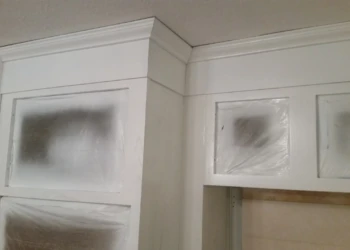 Close-up of white painted cabinetry corner with protective plastic on glass panels during cabinet refinishing painting in Westford