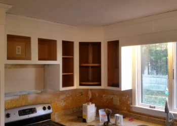 Partially refinished kitchen cabinets in corner with open wooden cabinet boxes, white painted frames, and countertop under window in Westford, MA