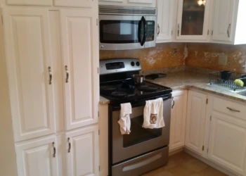 White kitchen cabinets installed around stainless steel oven and microwave with beige countertops and a window