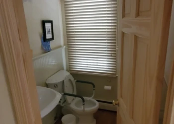 View into a small bathroom with a toilet, white sink, wooden floor and door, and window blinds in Weston MA