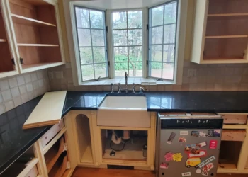 Open kitchen cabinets under black countertop around a farmhouse sink below a bay window in Weston MA kitchen renovation