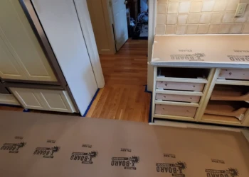 Partially refinished kitchen cabinets with drawer fronts and doors removed, hardwood floor covered for protection during work