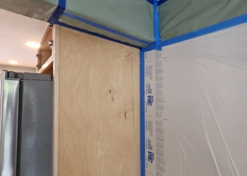 Bare wood cabinet side panel adjacent to refrigerator with surrounding area covered and taped for cabinet refinishing project in Windham NH