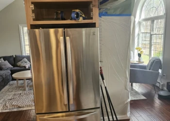 Wood kitchen cabinet above stainless steel fridge with painter's tape and protective plastic around adjacent walls in Windham NH