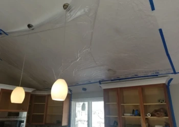 Ceiling drywall repair and painting in progress with plastic sheeting and lighting fixtures.
