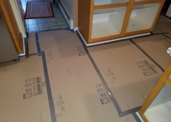 Protected kitchen floor with cardboard and tape near empty wooden cabinets during cabinet refinishing in Pelham, NH