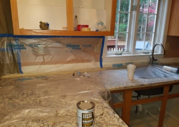 Kitchen counter and cabinets covered with protective plastic film and painter's tape during cabinet refinishing preparation in Pelham, NH