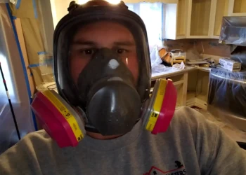 Worker wearing protective respirator mask and Finish Masters Builders sweatshirt in kitchen cabinet refinishing site in Pelham NH