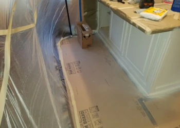 Protective plastic sheets and cardboard covering floor and counters around kitchen cabinets during cabinet refinishing in Pelham, NH