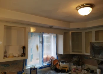 Unfinished kitchen cabinets without doors and protective plastic covering window and countertops during cabinet refinishing in Pelham, NH