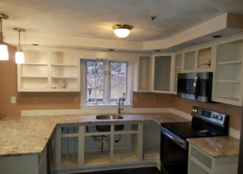 Kitchen with cabinets stripped of doors and hardware, granite countertops, window above sink ready for cabinet refinishing in Pelham NH