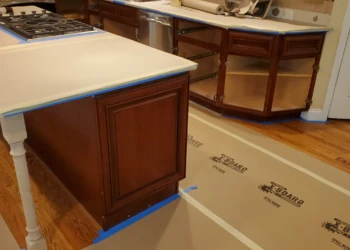 Kitchen cabinets with a dark wood finish and protected countertops and floors during cabinet refinishing in Windham, NH