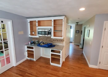 Kitchen area with upper and lower cabinet doors removed, showing bare wood interiors and countertop with painting supplies